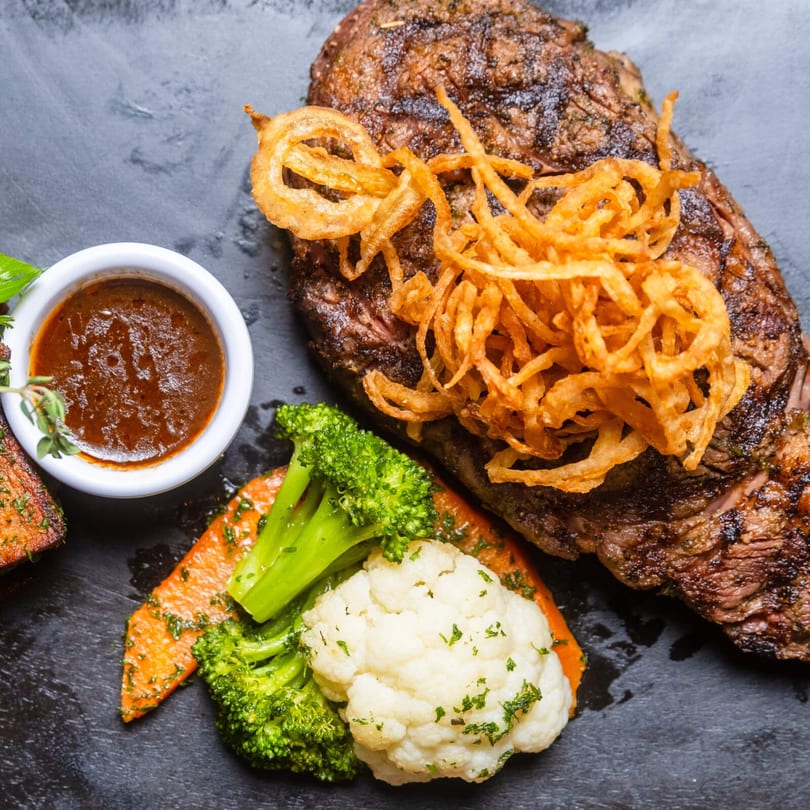 Grilled steak topped with crispy fried onions, served with broccoli, cauliflower, carrots, and a side of sauce.