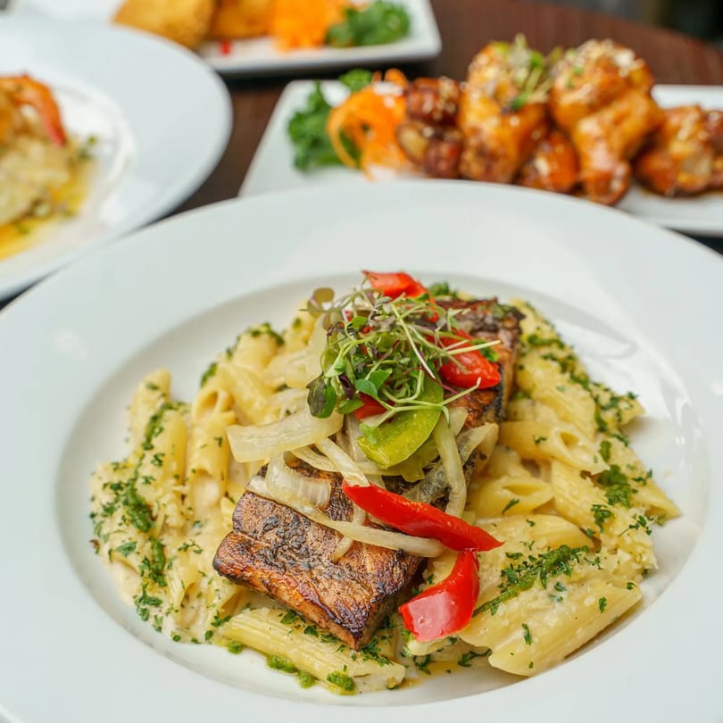 Grilled fish with bell peppers and onions on a bed of penne pasta, garnished with microgreens.