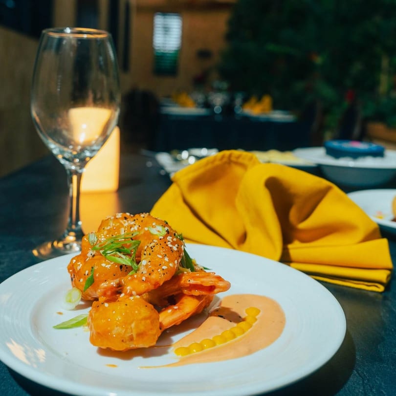 Plate of Caribbean shrimp with sauce, sesame seeds, and scallions, with a wine glass and napkin.