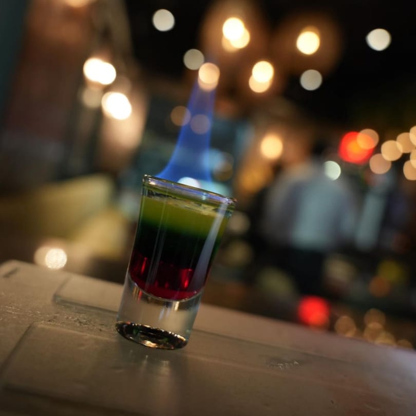 Flaming shot glass with layered red, black, and green liquids on a bar with bokeh lights.