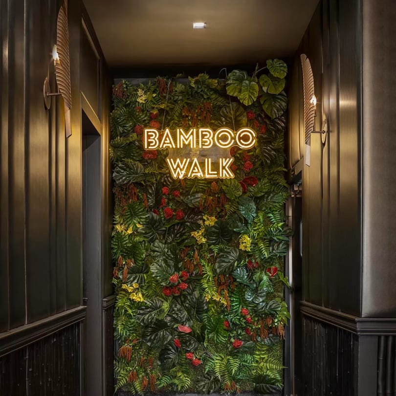 A neon "BAMBOO WALK" sign glows against a lush green wall with red and yellow flowers.