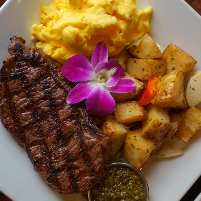 Grilled steak, scrambled eggs, and seasoned potatoes with a purple orchid garnish on a white plate.