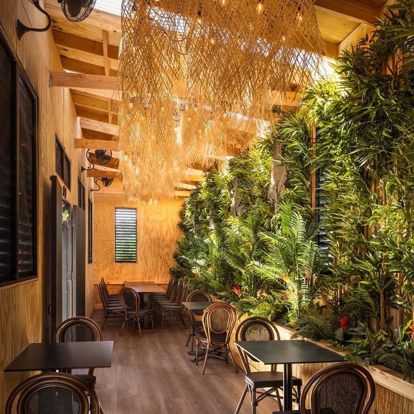 Bamboo Walk Caribbean Restaurant interior with wooden walls, lush green plants, and rattan light fixtures.