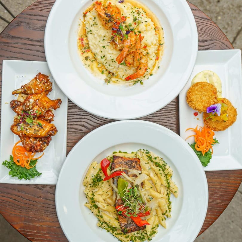 A table laden with Caribbean dishes: shrimp and grits, jerk chicken wings, crab cakes, and salmon pasta.
