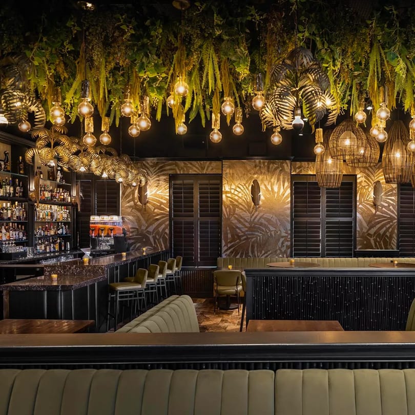 Bamboo Walk Caribbean Restaurant interior with a bar, green velvet seating, and tropical decor with hanging plants and lights.