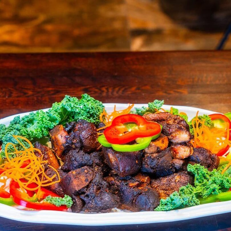 A white platter of dark, richly seasoned Caribbean jerk chicken, garnished with fresh kale and colorful bell peppers.