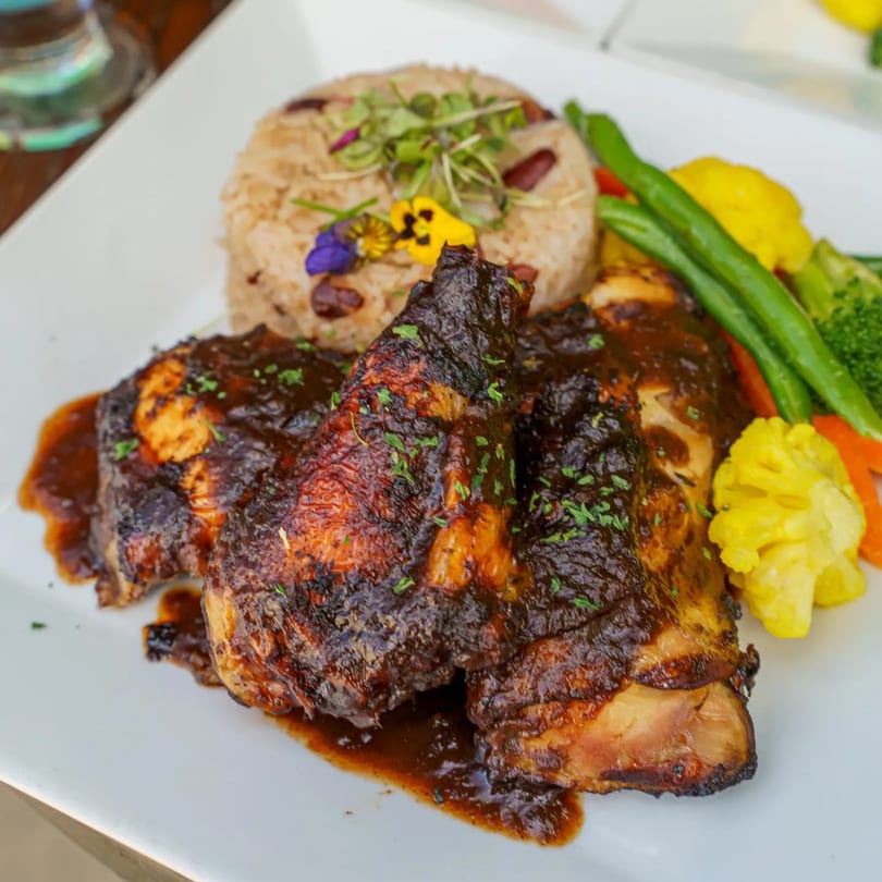 A plate of Caribbean jerk chicken with rice and peas, garnished with edible flowers and mixed vegetables.