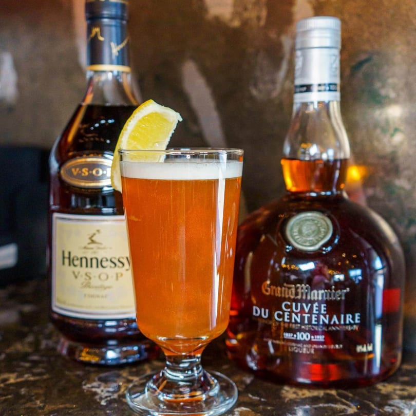 A Caribbean cocktail with a lemon slice, flanked by bottles of Hennessy and Grand Marnier.