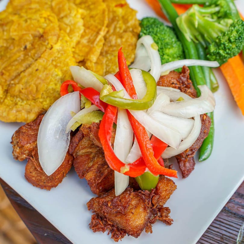A Caribbean dish with fried plantains, fried fish topped with peppers and onions, and steamed vegetables.