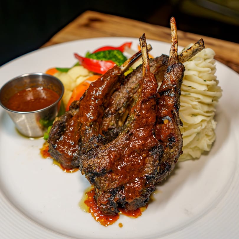 Grilled lamb chops with sauce, mashed potatoes, and mixed vegetables on a white plate.