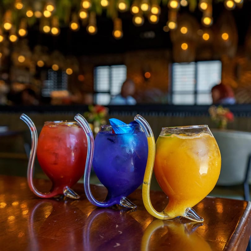 Three colorful Caribbean cocktails in unique glasses with built-in straws on a wooden table.