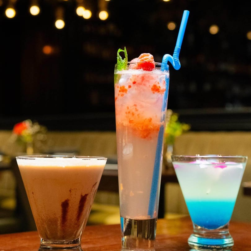 Three vibrant Caribbean drinks, including a pink cocktail with a blue straw, on a wooden table.