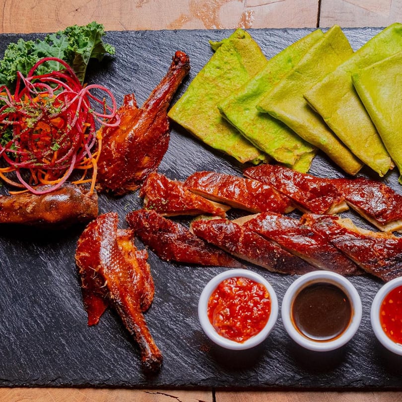 Roasted duck, whole and sliced, with green pancakes, kale, and three dipping sauces on a dark platter.