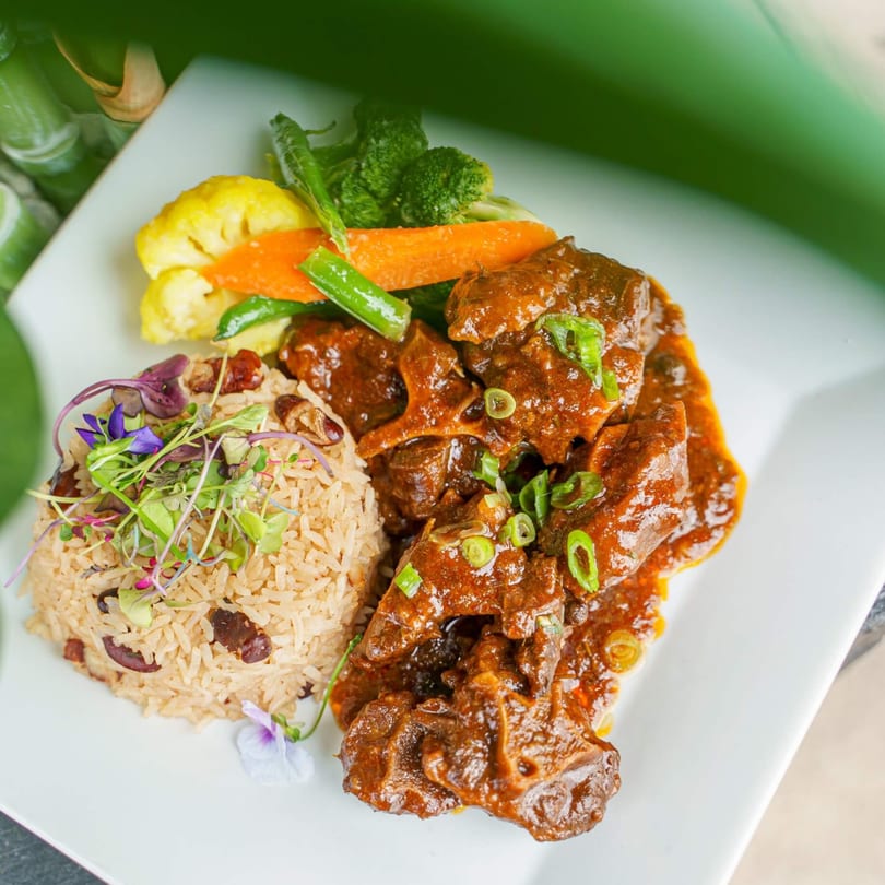 Oxtail stew with rice and peas, garnished with microgreens and edible flowers, served with steamed vegetables.