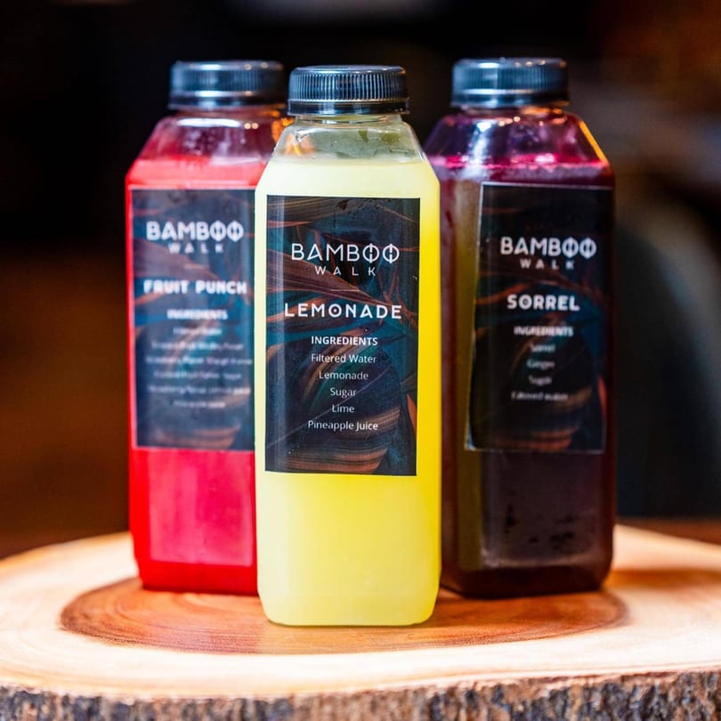 Three Bamboo Walk bottled drinks: red fruit punch, yellow lemonade, and dark sorrel, on a wooden surface.