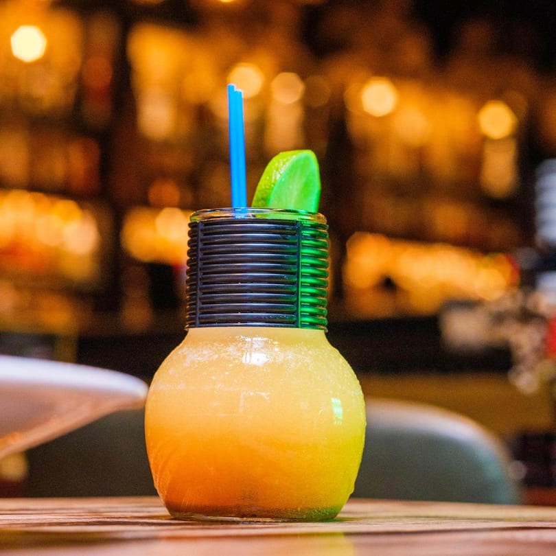 A vibrant orange Caribbean drink in a unique glass with a blue straw and lime wedge.