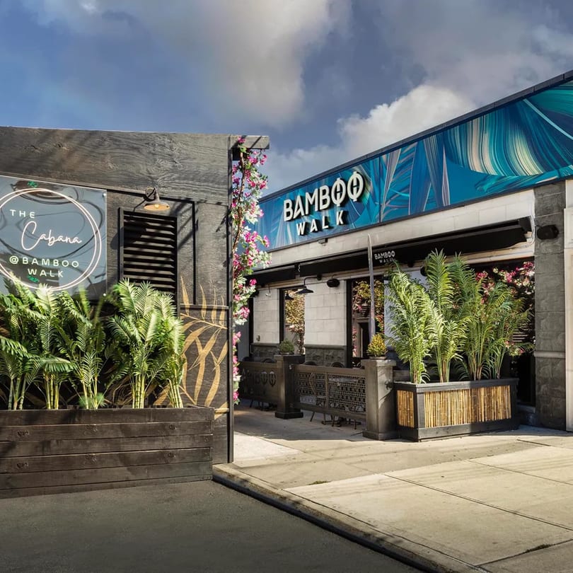 Exterior of Bamboo Walk Caribbean Restaurant with lush greenery, a blue facade, and outdoor seating.