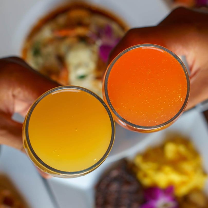 Two hands holding glasses of orange and yellow juice, with Caribbean food blurred in the background.